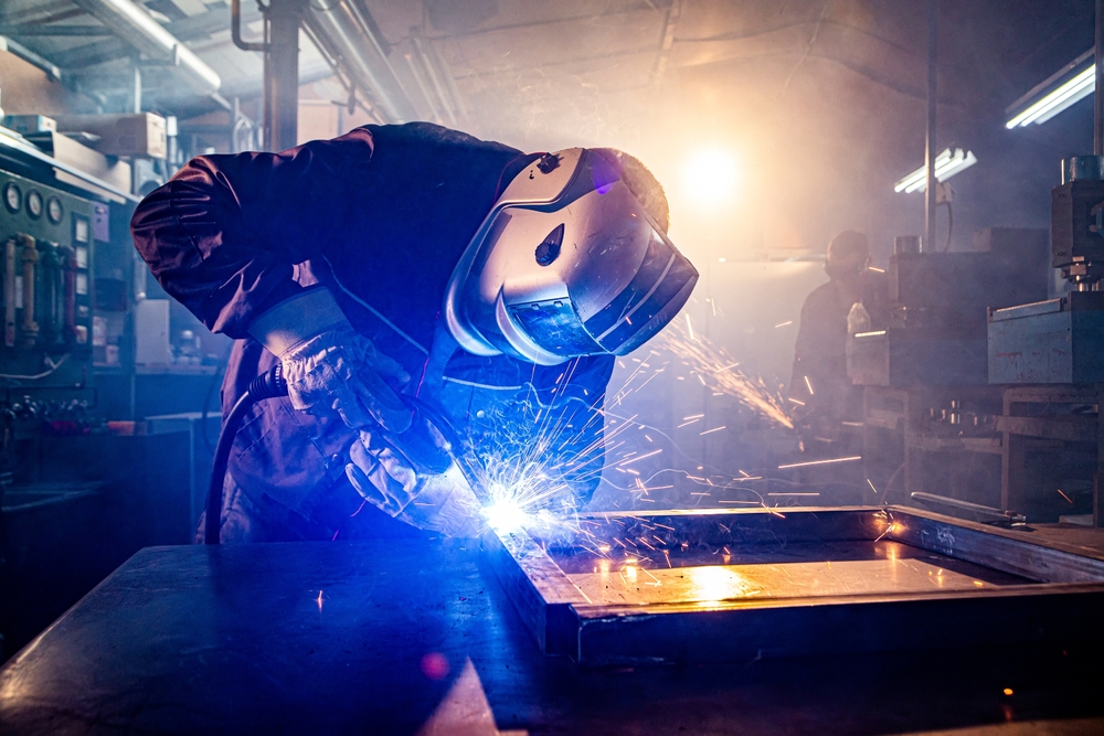 A welder in protective gear creates sparks while working on a metal frame in a dimly lit workshop, highlighting his focused craftsmanship